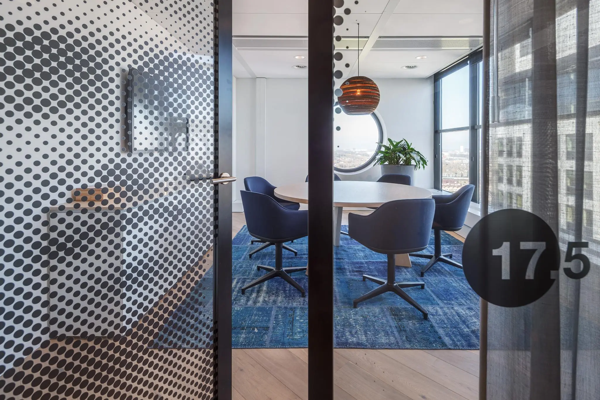 Moderne vergaderruimte in Rivium Quadrant met ronde tafel, blauwe stoelen en uitzicht door een rond raam.